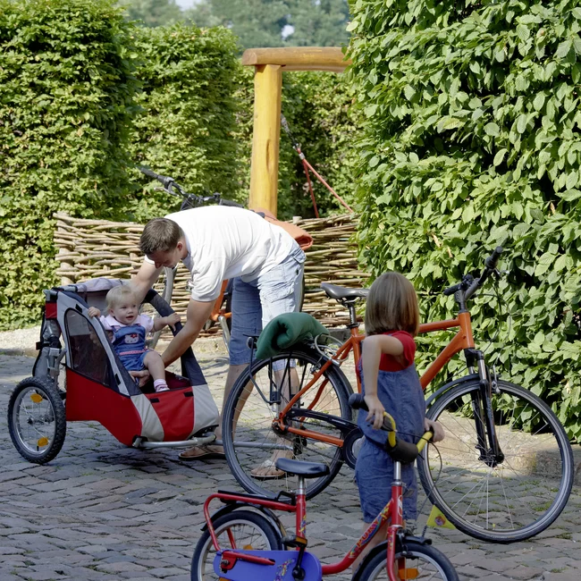 Vater überprüft kleines Kind im Fahrradanhänger, anderes Kind mit Laufrad daneben