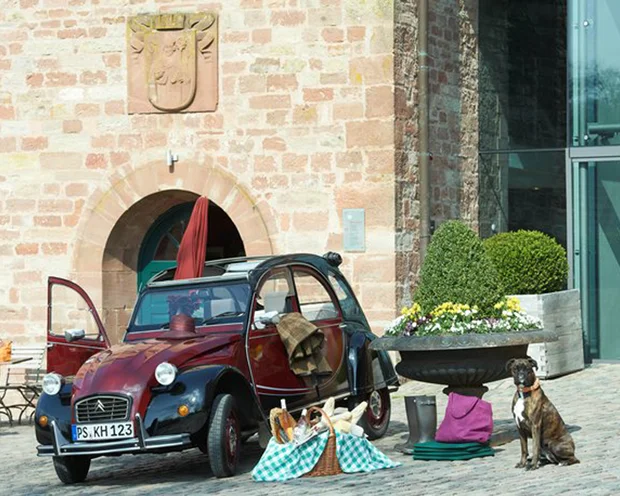 Rotes und schwarzes Vintage-Auto mit Picknickkorb und sitzendem Hund vor altem Gebäude