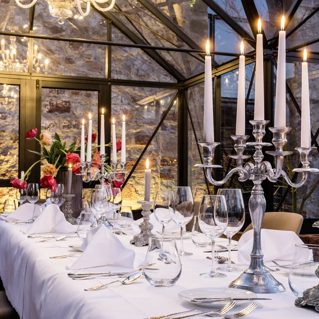 Eleganter Tisch mit Kerzenständern, Kerzen, Gläsern und Blumen in einem Glasgewächshaus