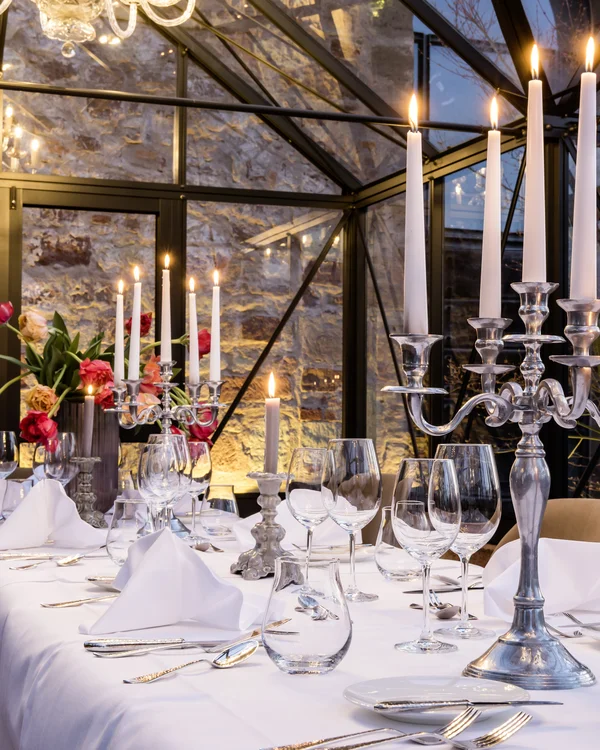 Eleganter Tisch mit Kerzenständern, Kerzen, Gläsern und Blumen in einem Glasgewächshaus