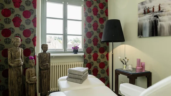 Behandlungsraum mit Liege, Handtüchern und asiatischen Holzfiguren unter Fenster