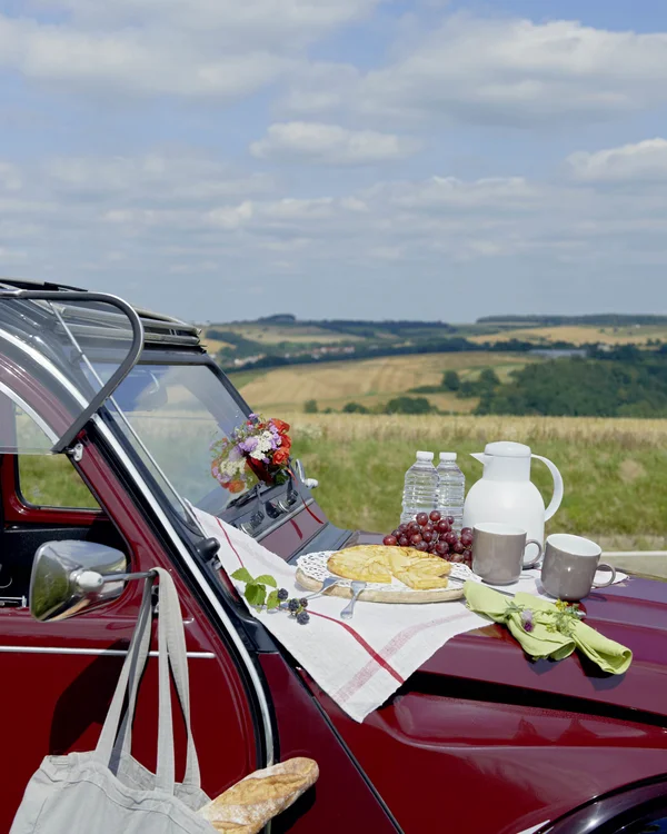 Picknick mit Kuchen und Getränken auf der Motorhaube eines roten Autos in der Natur