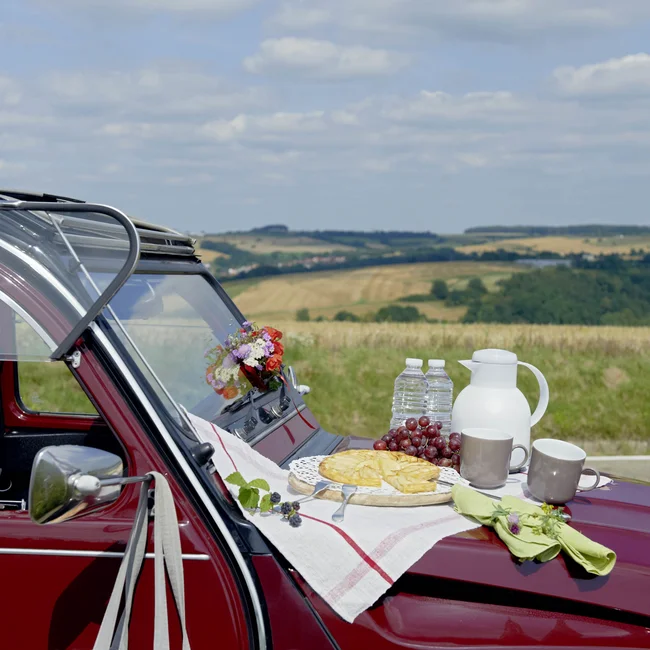 Picknick mit Kuchen und Getränken auf der Motorhaube eines roten Autos in der Natur