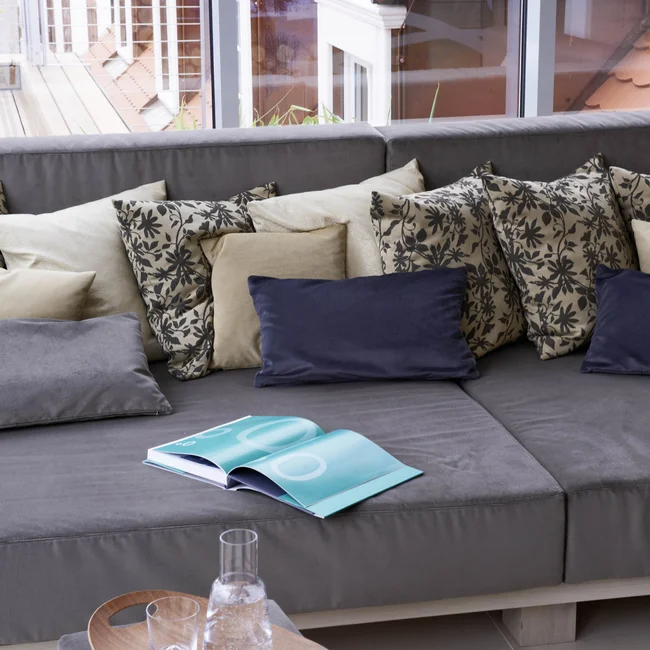 Graues Sofa mit gemusterten Kissen vor großem Fenster mit Dachansicht