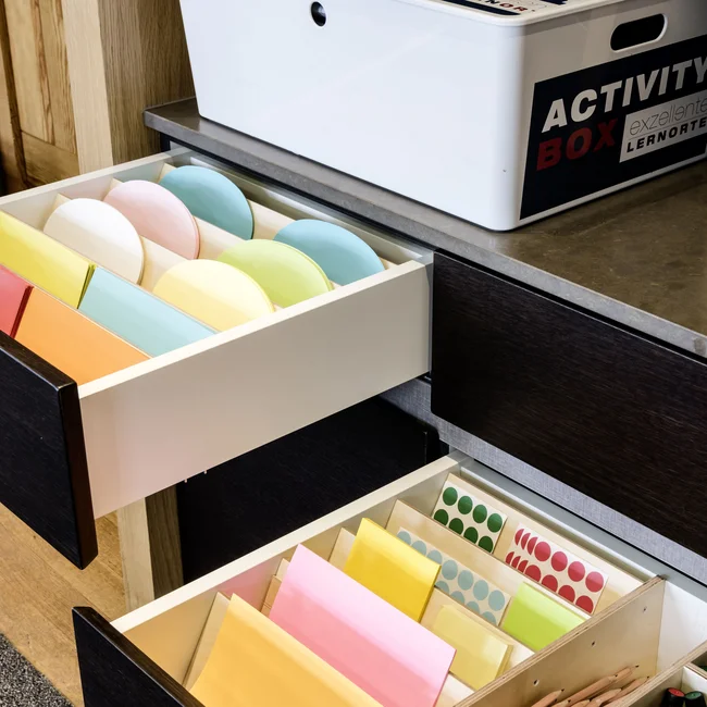 Offene Schubladen mit buntem Papier und Stiften neben einer weißen Activity Box