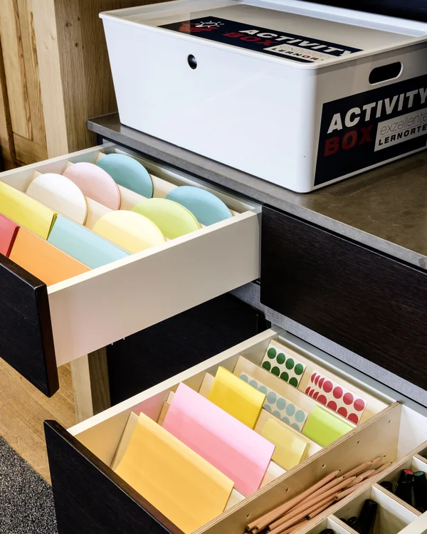 Offene Schubladen mit buntem Papier und Stiften neben einer weißen Activity Box