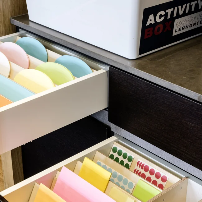 Offene Schubladen mit buntem Papier und Stiften neben einer weißen Activity Box
