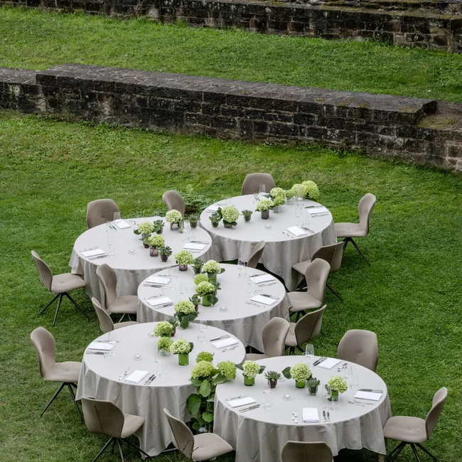 Mehrere gedeckte Tische mit Blumen auf grünem Gras vor einer Steinmauer