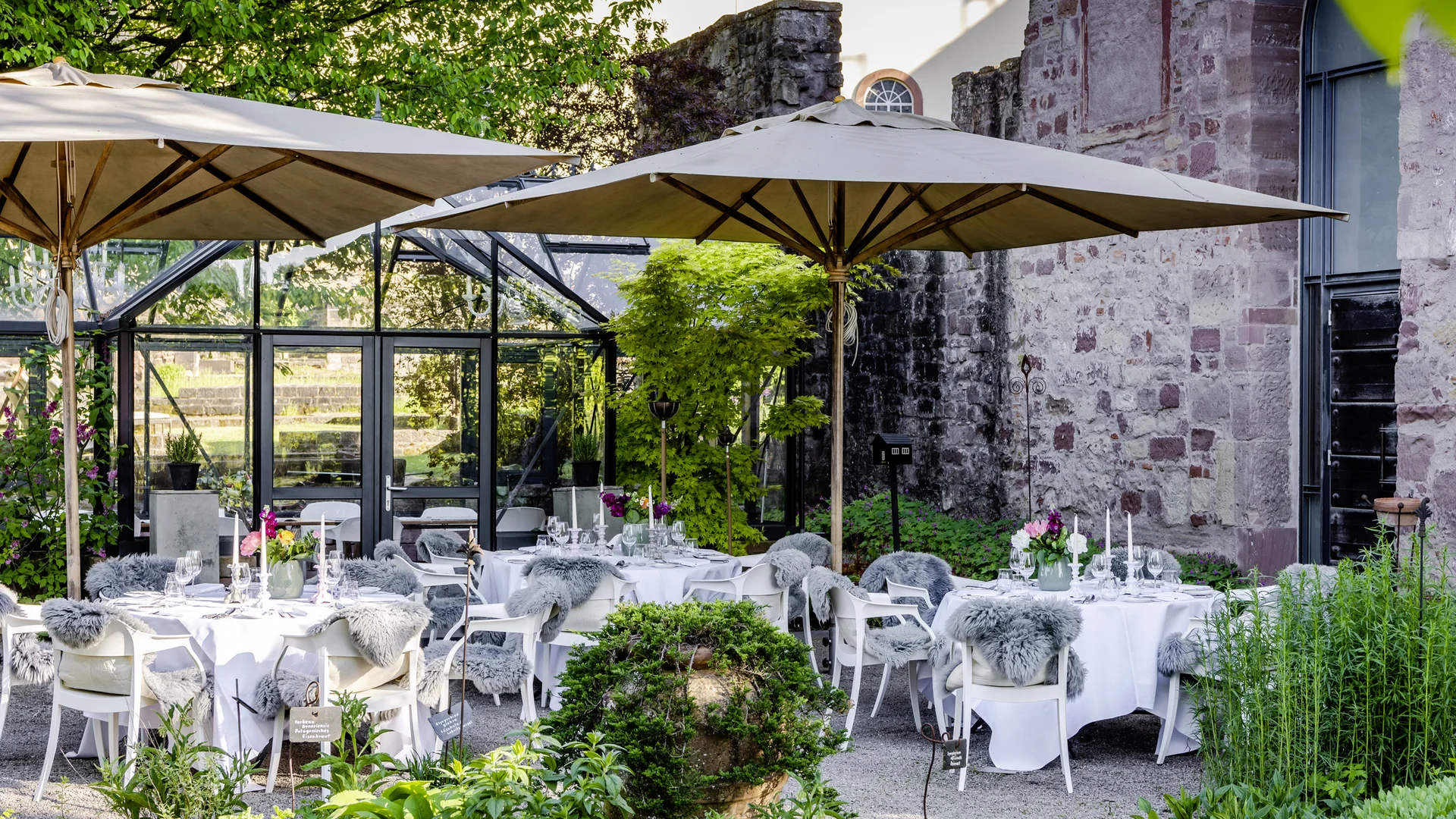 Was wäre Feiern ohne köstliches Essen und passende Drinks? Elegante Außenterrasse mit Tischen unter Sonnenschirmen und dekorativer Bepflanzung