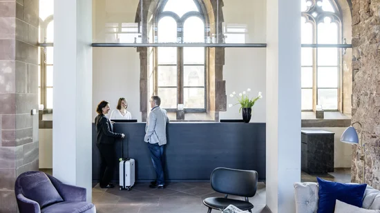 Hotelrezeption mit Gästen und Mitarbeiterin in einem Gebäude mit gotischen Fenstern