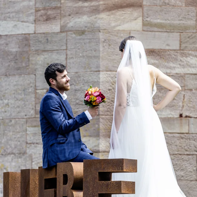 Bräutigam kniet vor Braut mit Blumenstrauß neben LIEBE-Skulptur